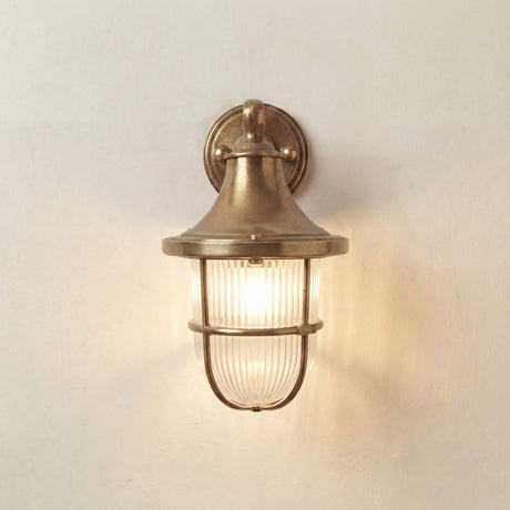 A vintage-style nautical wall sconce with a warm brass finish and a ribbed glass shade, illuminated against a textured off-white wall.