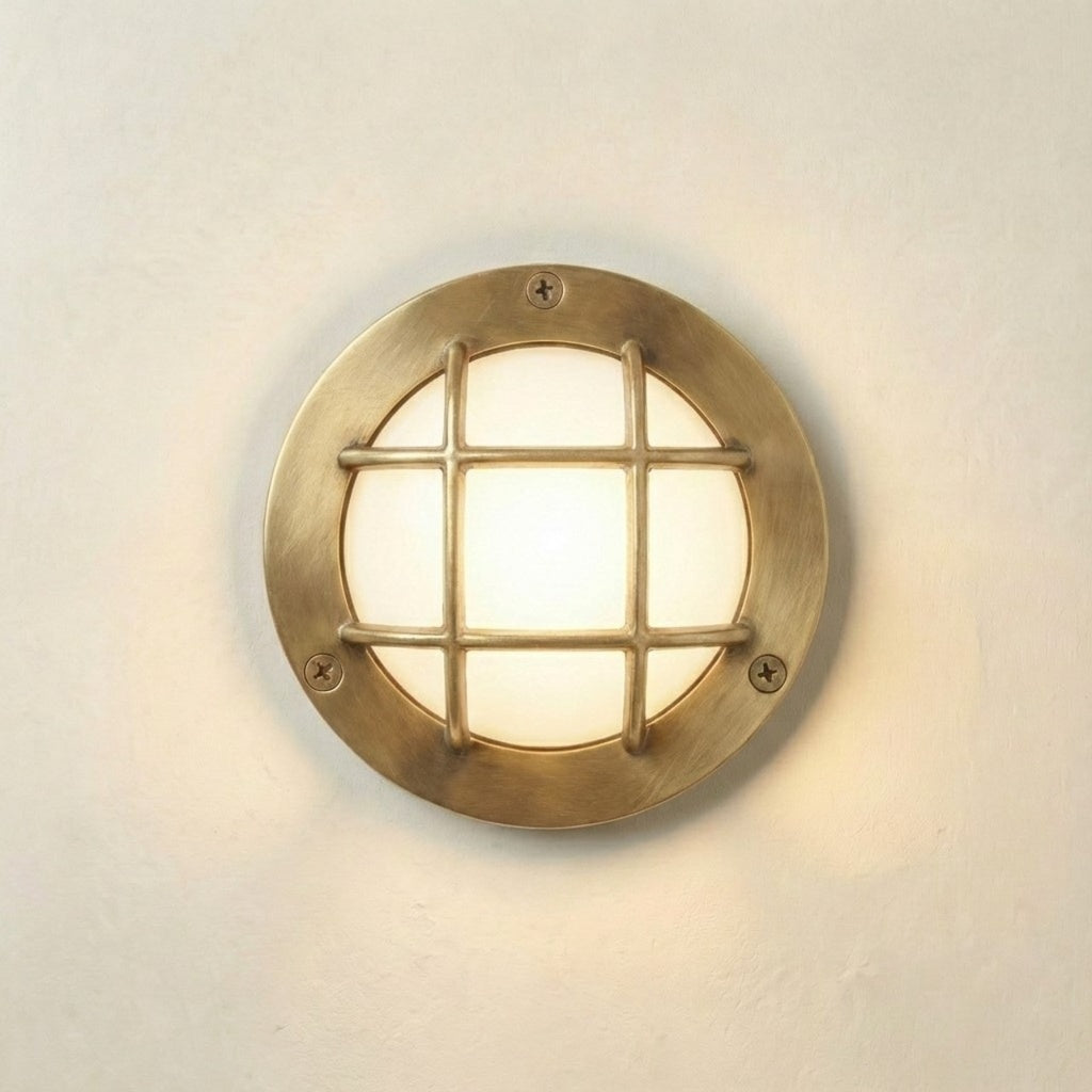 A round nautical-style wall light fixture with a brass-finished metal frame and a grid grille over a glowing frosted glass center, mounted on a plain off-white wall.