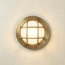 A round nautical-style wall light fixture with a brass-finished metal frame and a grid grille over a glowing frosted glass center, mounted on a plain off-white wall.