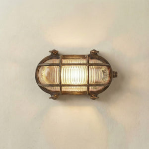 A vintage-style nautical wall sconce with a bronze-finished metal frame and ribbed clear glass panels, illuminated against a plain off-white wall.