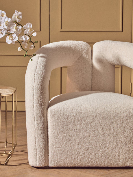 Beige textured chair with a gold side table and white flowers against a beige wall.