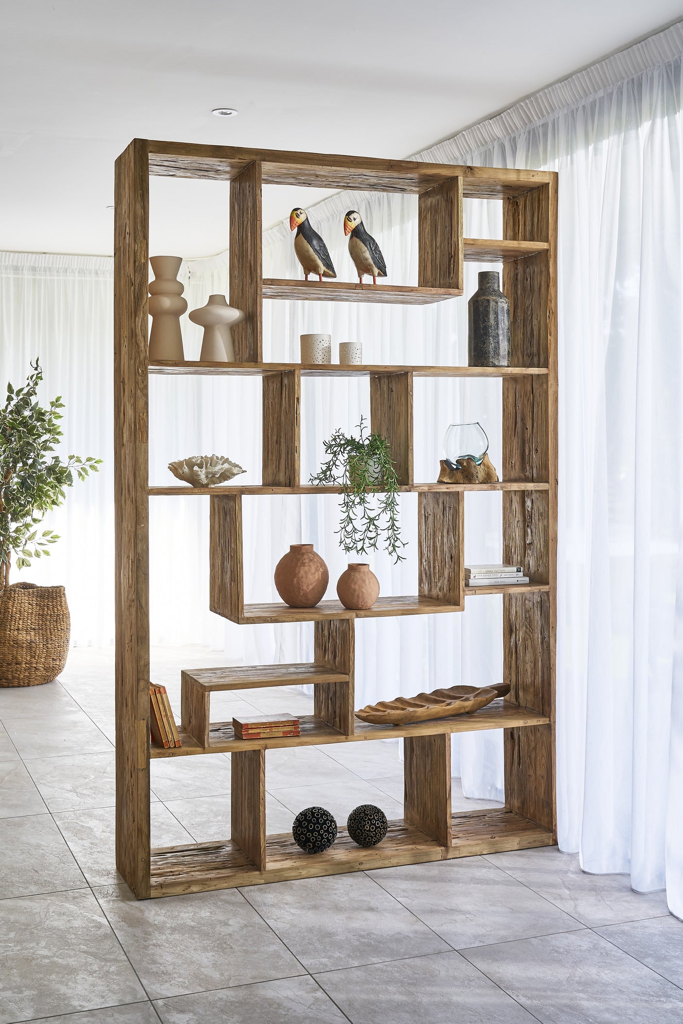 Wooden bookshelf with decorative items in a room with white curtains and a plant.