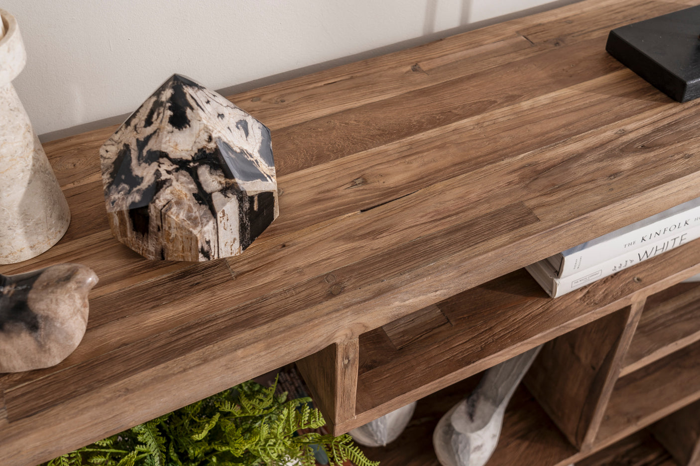 A close-up of a rustic wooden console table with open cubbies, styled with a black and white marbled crystal, a ceramic vase, books, and greenery.
