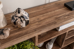 A close-up of a rustic wooden console table with open cubbies, styled with a black and white marbled crystal, a ceramic vase, books, and greenery.