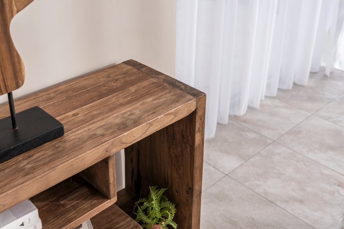 A close-up of a rustic brown wooden console table or shelf in a home setting, with a small green plant inside the open lower compartment and a white curtain and tiled floor in the background.