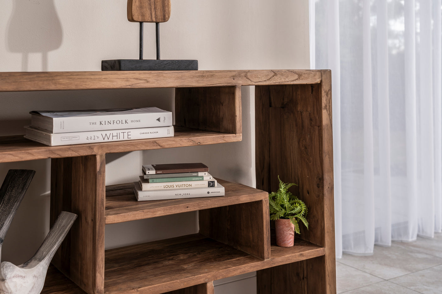 A rustic wooden open bookshelf styled with books, a small potted fern, and a decorative object on top, set against a neutral wall with sheer white curtains.