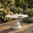 A round outdoor dining table with a sculptural pedestal base, set on a stone patio in a lush garden with flowers and plants in the background.