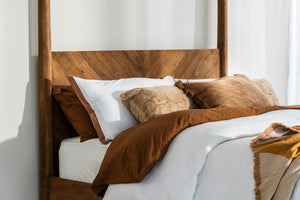 The Ashcombe four poster bed from the side, showing the honey-toned reclaimed teak headboard and post with layered bedding.