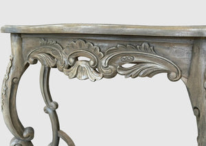 Close-up of an ornate carved console table leg and apron with a distressed gray wood finish, showing detailed scrollwork and cabriole-style curves against a light gray background.