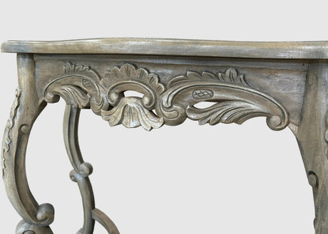 Close-up of an ornate carved console table leg and apron with a distressed gray wood finish, showing detailed scrollwork and cabriole-style curves against a light gray background.