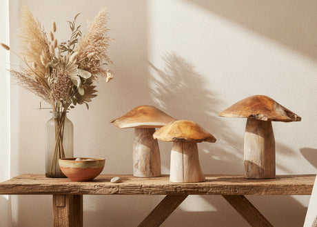 Three wooden mushroom sculptures on a wooden table with a vase of dried plants in the background.