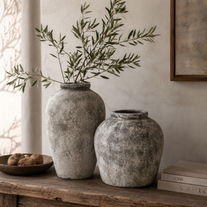 Two rustic stone vases with green branches on a wooden surface against a neutral wall.