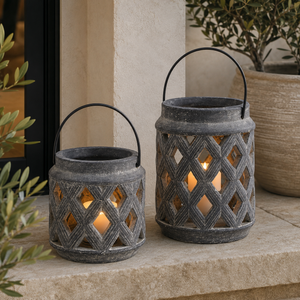 Two decorative metal lanterns with black wire handles sit on a stone ledge outdoors. Each lantern has a cutout lattice pattern and a warm glowing candlelight insert.
