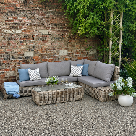 Outdoor furniture set arranged in a corner, consisting of a sofa with cushions and a coffee table, made of wicker, on a patio with a brick wall in the background.