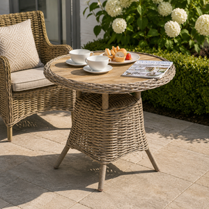 Wicker outdoor table with tea set and bread, surrounded by chairs and flowers.