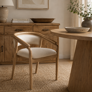 A light oak wood armchair with a cream upholstered seat and back cushion, styled in a warm, rustic room next to a matching wood table.