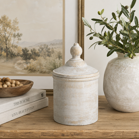 Antique white canister with lid on a wooden surface with a vase and greenery, books and artwork