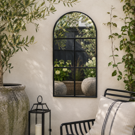 Arched black metal wall mirror with a six-pane grid design, mounted on a textured cream stucco wall in a garden patio setting.