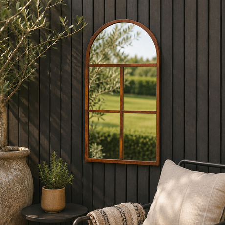 An arched wooden-framed mirror with a six-pane grid design is mounted on a dark charcoal wood-paneled wall. The mirror reflects a green outdoor lawn and trees.