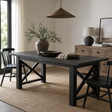 A dark charcoal black farmhouse dining table with X-shaped legs, styled in a warm, neutral-toned dining room with black chairs, a wooden credenza, and a woven pendant light.