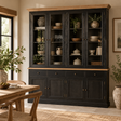 A large black wooden hutch with glass upper doors and a light wood top, styled in a dining room with dishes, plants, and a wooden table nearby.