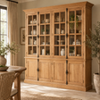 A large light oak wood china cabinet with glass-front upper doors and panelled lower doors, styled in a warm, sunlit dining room.