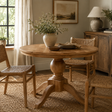 Round wooden pedestal dining table in a warm, rustic room setting with woven chairs, a vase of greenery, and neutral decor.