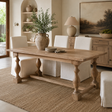 A rustic wooden dining table with turned legs and a rectangular top, styled in a warm, neutral-toned dining room with white upholstered chairs, a woven rug, and a large ceramic vase on top.