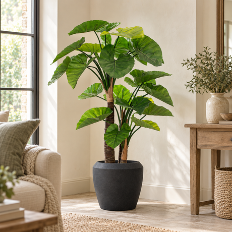 Artificial Alocasia Tree in a black planter in a living room, beside a wooden side table, an armchair and a woven basket