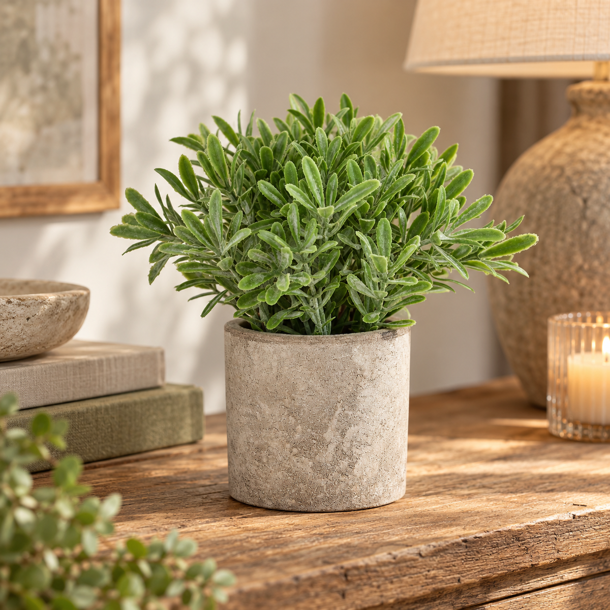 Artificial Buxus Plant in a stone-effect pot on a wooden sideboard beside books, a lit candle and a table lamp