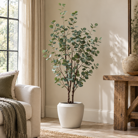 Artificial Eucalyptus Tree with White Planter in a living room, beside an armchair with a knitted throw and a rustic wooden console