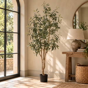 A tall artificial olive tree in a black pot, styled in a bright room next to a window, console table, and lamp.