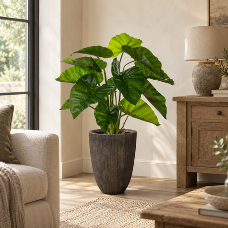 Artificial Taro Plant in a tall ridged graphite planter in a living room, beside a cream armchair and a wooden cabinet with a stone lamp