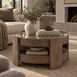 A round wooden coffee table with a glass top and open lower shelf, styled in a living room with books, a vase, and bowls.