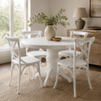 A round white pedestal dining table with four matching white chairs in a bright, neutral-toned dining room. A beige ceramic pitcher with greenery and a small wooden bowl sit on the tabletop.