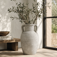 Gray textured vase with green branches on a wooden surface near a window.