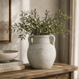 Decorative vase with greenery on a wooden surface with books and a framed picture in the background.