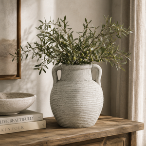 Decorative vase with greenery on a wooden surface with books and a framed picture in the background.