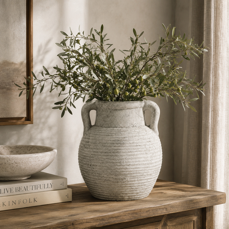 Decorative vase with greenery on a wooden surface with books and a framed picture in the background.