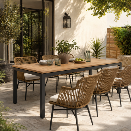 Outdoor dining set with a rectangular wooden table and woven wicker chairs on a patio, surrounded by plants and a light-colored stucco wall.