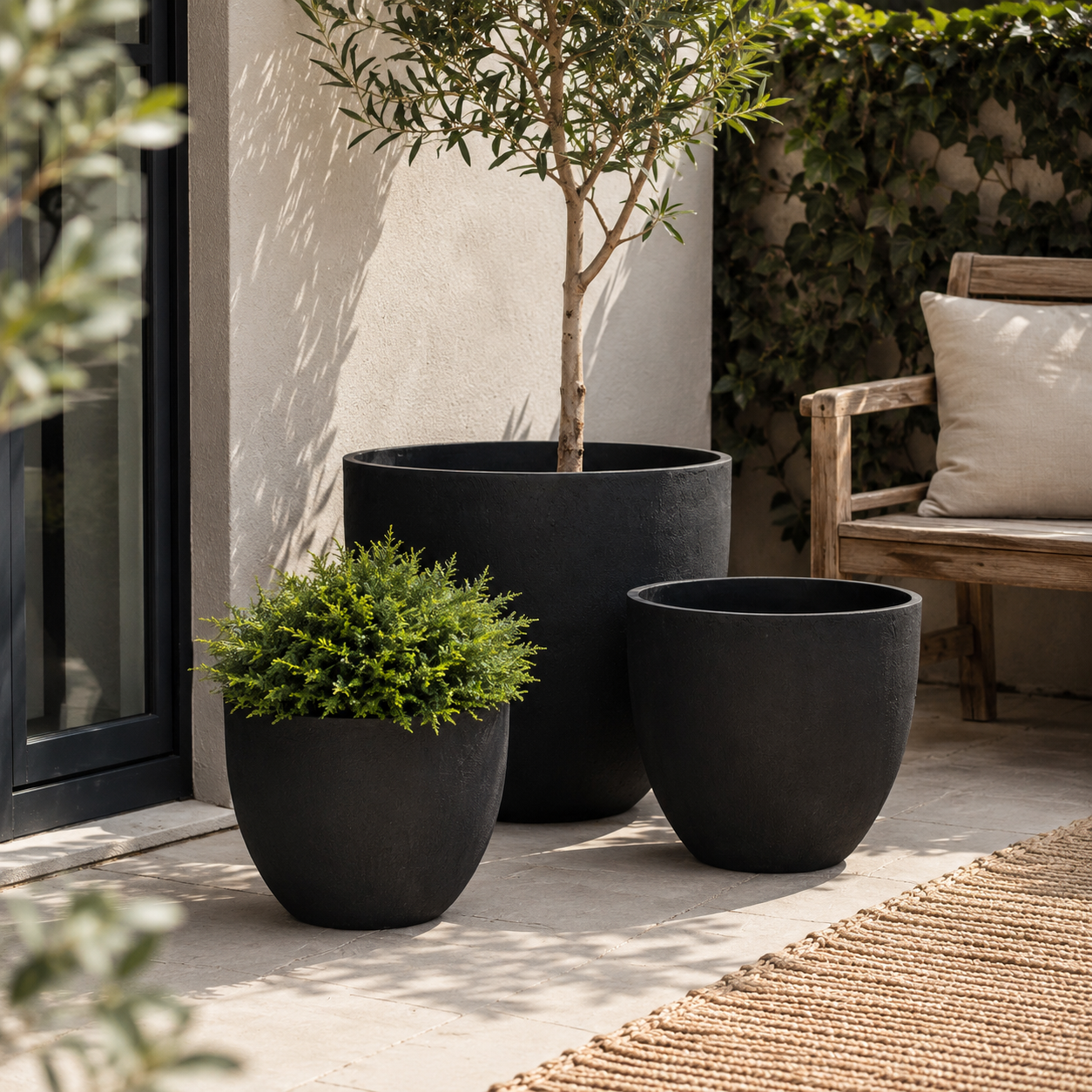 Three matte black outdoor planters in graduated sizes on a patio, with a small green shrub in the front left planter, a tall tree in the back center planter, and an empty planter beside it. A wooden bench with a cream pillow and a leafy wall are visible in the background.