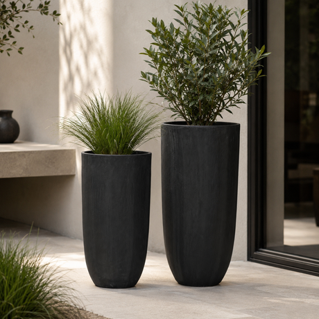 Two tall matte black planter pots with rounded tapered shapes, one holding a green ornamental grass plant and the other a leafy shrub, set on a light stone patio in front of a beige stucco wall.