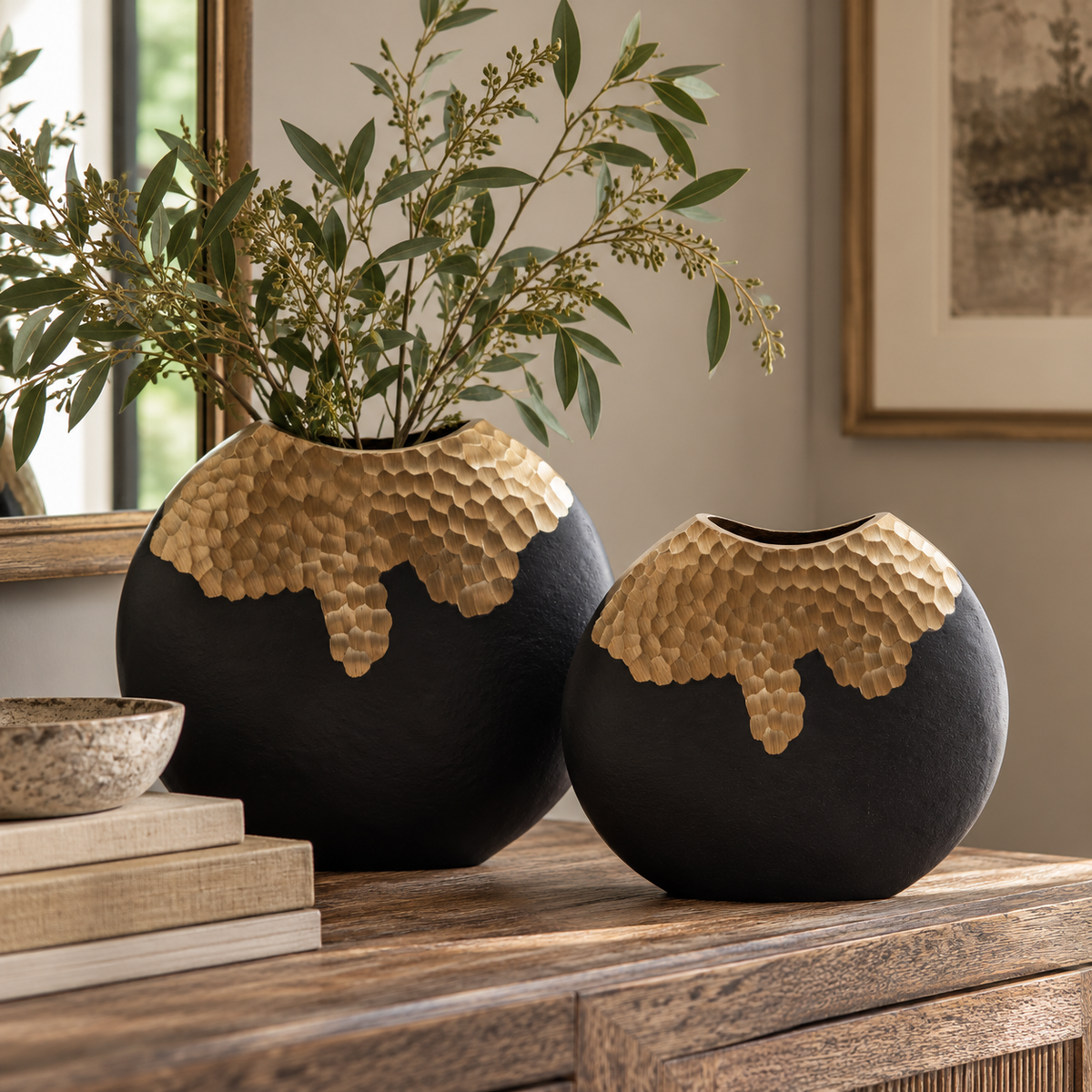 Two Aurum Round Black and Gold Vases in different sizes on a wooden sideboard, the larger holding olive branches, beside a stack of books