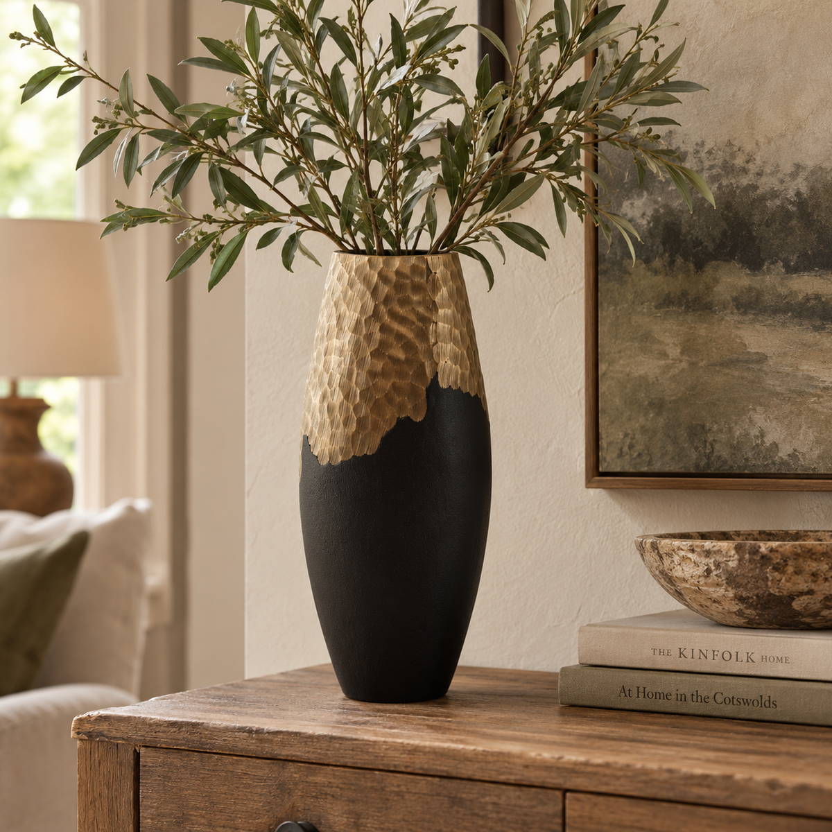Tall Aurum black and gold vase filled with olive branches on a wooden chest of drawers, beside books and a rustic wooden bowl