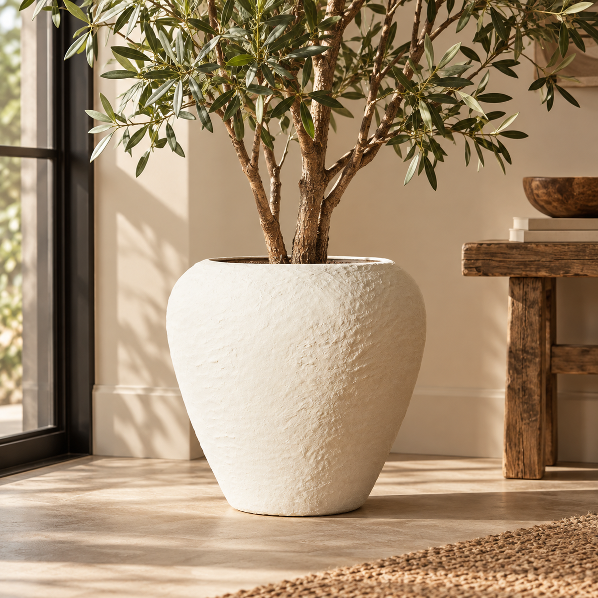 Avara Cotton Mache Large White Planter holding an olive tree on a pale stone floor by a window, beside a rustic wooden side table and a jute rug