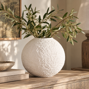 Avara Cotton Mache Small Round White Vase holding olive branches on a wooden sideboard, beside a framed print and a stoneware bowl