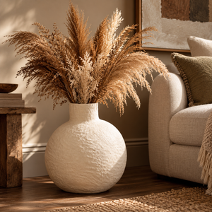 Avara Large Cream Cotton Mache Vase filled with dried pampas grass on a wooden floor beside a cream armchair and a rustic side table
