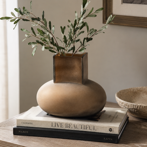 A modern ceramic vase with a matte beige rounded base and a square metallic bronze top, holding green olive branches, styled on a stack of books on a wooden table.
