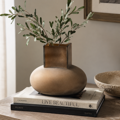 A modern ceramic vase with a matte beige rounded base and a square metallic bronze top, holding green olive branches, styled on a stack of books on a wooden table.