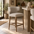 A modern bar stool with a textured light beige upholstered seat and backrest, supported by four tapered natural wood legs, shown in a bright, sunlit kitchen setting.